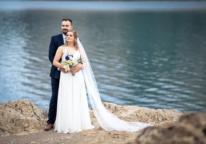 Wedding Portrait by the Minnewanka Lake. Groom hugs the bride from behind and they both look at the same direction.