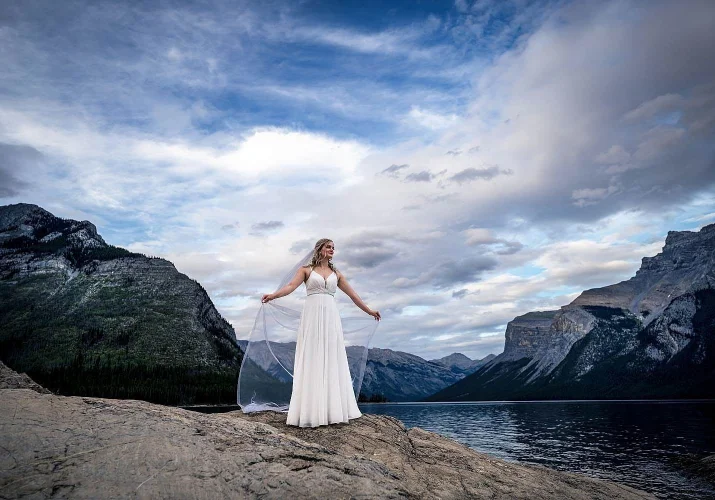 Amazing bride's portrait at Minnewanka Lake captured by Banff Wedding Photographers. Bride holds her veil and look at the lake.