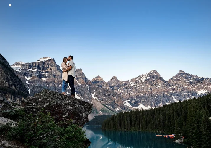 Newly engaged couple in love hugging during their engagement session at Moraine Lake. They stand at the edge ot the rock against tremendous mountains and lake/