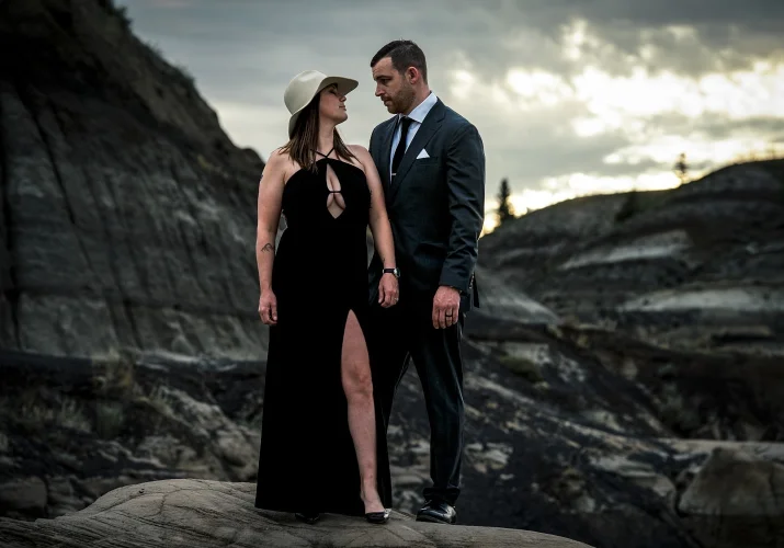 Married couple during after-wedding session. lady wears log black dress and a hat and Gentleman wears suit. They loos at each other with love.
