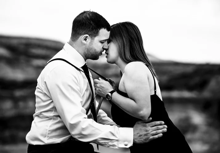 Black and white photograph of couple in love. Girl pulls her husbands suspenders and kisses him.