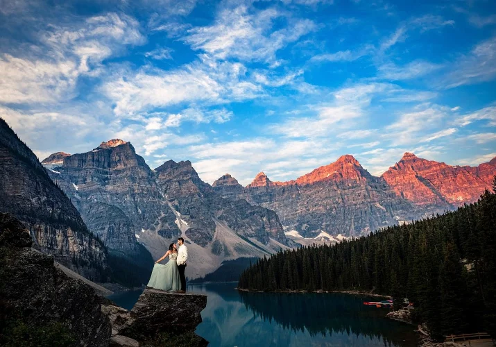 Engaged couple stand on the huge rock at Moraine Lake surrounded by the Canadian Rockies. Lady plays with her dress.