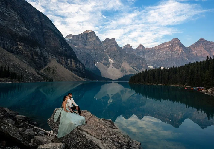 Young and handsome couple in love sit at the rock against huge Canadian Rocky Mountains at Moraine Lake.