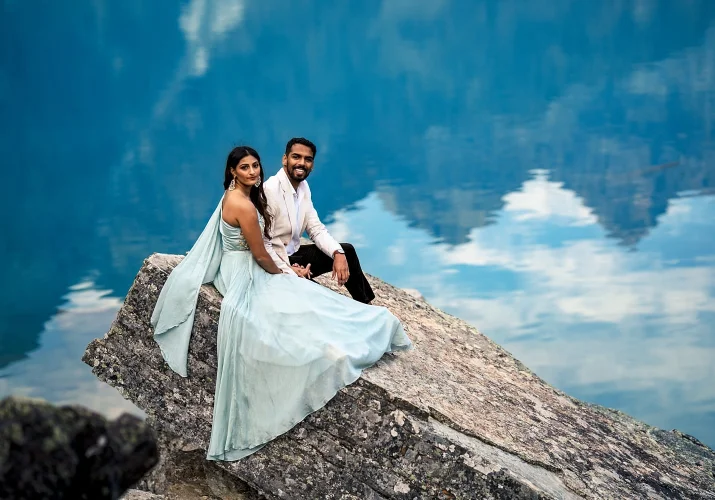 lady in a long, blue dress and her fiancee sit at the rock by the Moraine Lake in Banff.