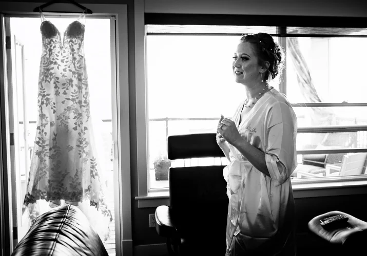Photograph of the bride wearing a robe and a wedding dress hanging on the window.