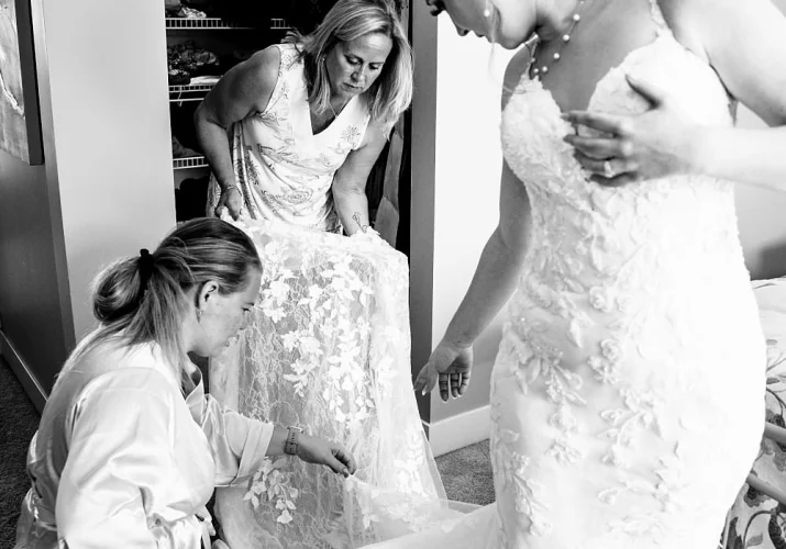 Bride's Mother and Bridesmaid help the bride with the wedding dress. Brides points out on the buttons on her dress.