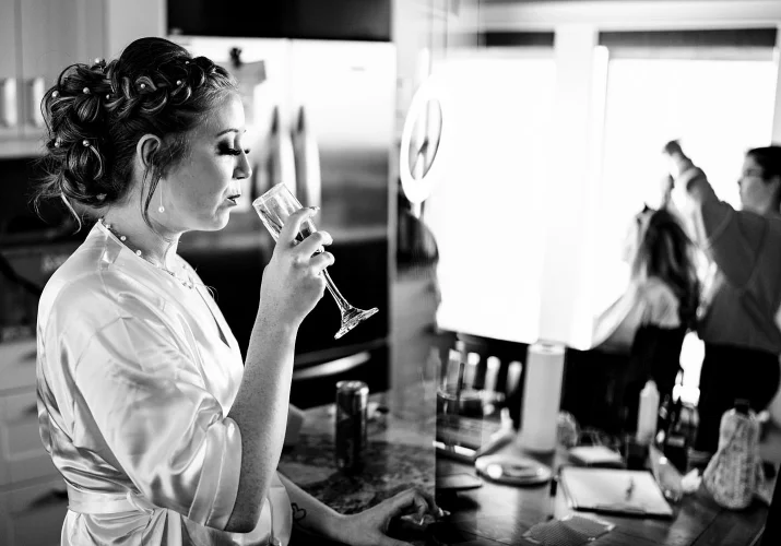 Photograph of the bride wearing a robe and drinking a glass of champagne before wedding ceremony. On the background bridesmaid and makeup artist.