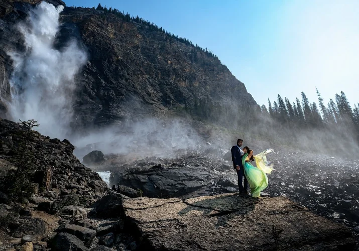 Engagement photograph of couple in a hug at Takakkaw Falls. Wind is blowing the girl yellow dress.