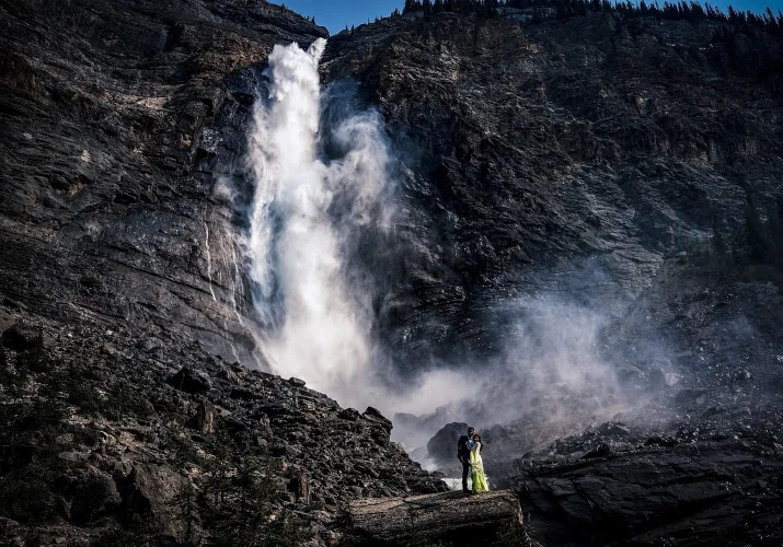 Huge Takakkaw Falls with tiny engaged people hugging each other.