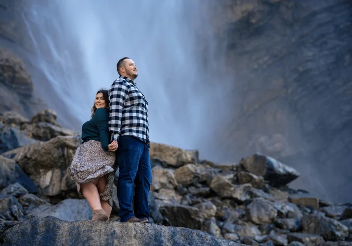 Engaged couple standing back to back and holding hands against breathtaking Takakkaw Falls in Banff.