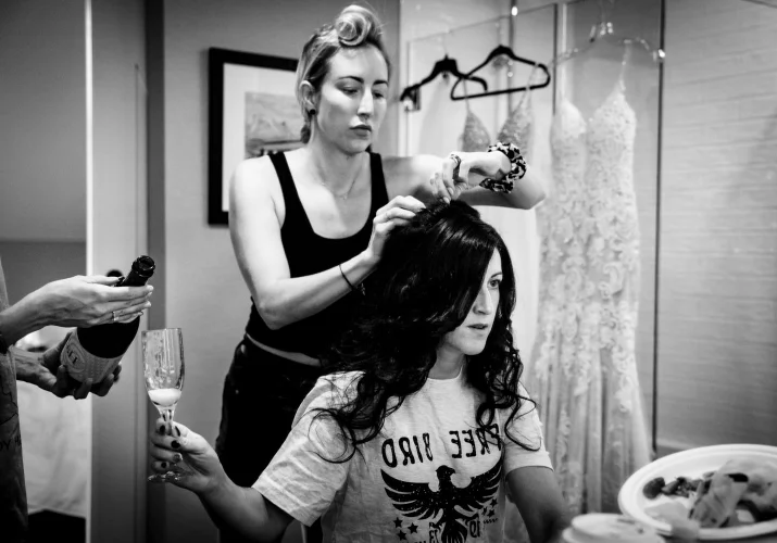 Bride is getting her hair done while her friend fill her glass with champagne