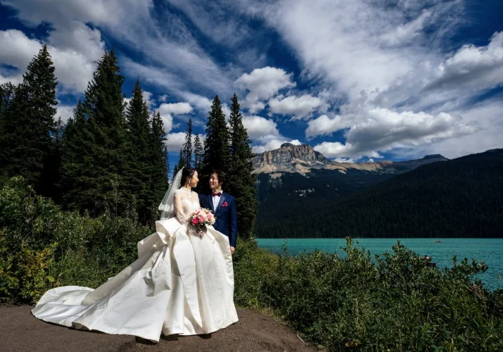 Newlyweds portrait at Emerald Lake . Behind the couple tall trees , lake and the mountains. Above their heads interesting sky with moving clouds,