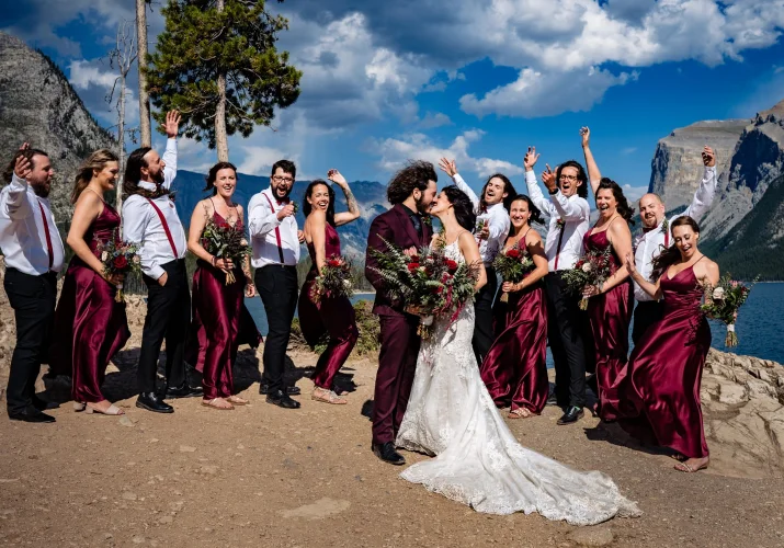 Bridal Party at Minnewanka Lake. Newlyweds share a kiss while bridal party cheer them.