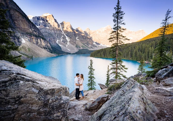 Engagement young couple posing to Banff Wedding Photographer at amazing location of Moraine Lake. They are in a hug, facing each other. Behind them beautiful lake and huge mountains