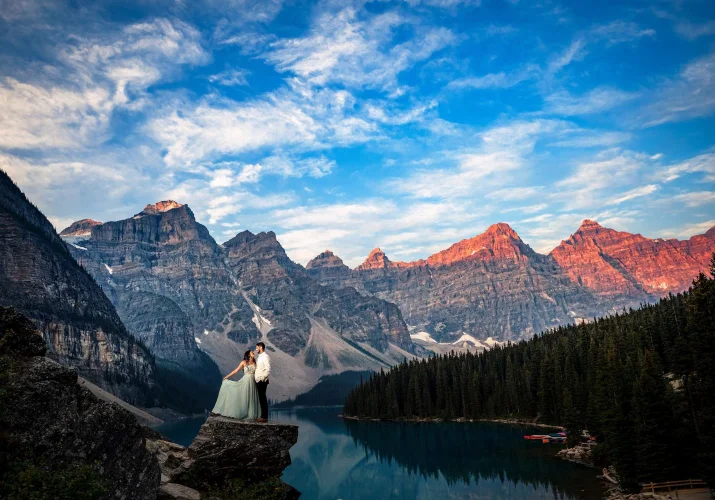 Bride and groom on the rock by Moraine Lake. Bride plays with her wedding dress while the groom look at her. Behind them tremendous Canadian Rockies.