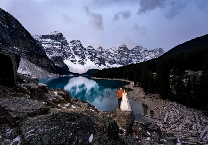 Incredible Banff Wedding Portrait at Moraine Lake of newlyweds standing at the stone. Behind them turquoise lake and Canadian Rocky Mountains covered with snow.