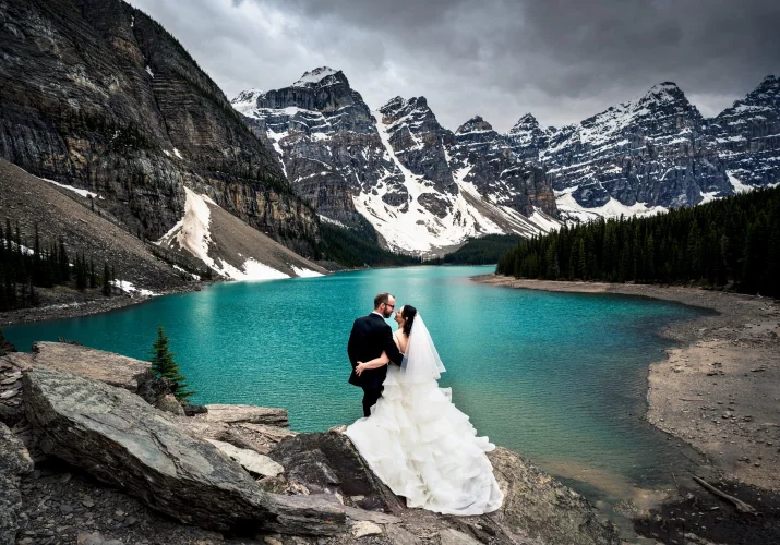 Newlyweds standing by Moraine Lake and admire the beauty of turquoise Moraine Lake and tremendous mountains covered with snow. Moment captured by the best Banff Wedding Photographer.