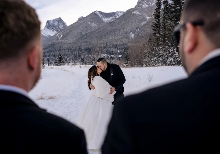 Bride and groom share a kiss while guests are watching them. Photo taken in Banff with the Rocky Mountains on the background.